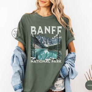 Może przedstawiać: Zielona koszulka w kolorze mchu z grafiką Parku Narodowego Banff. Projekt zawiera napis "BANFF" nad malowniczą ilustracją jeziora, g&oacute;r i drzew. Poniżej ilustracji znajdują się słowa "NATIONAL PARK".