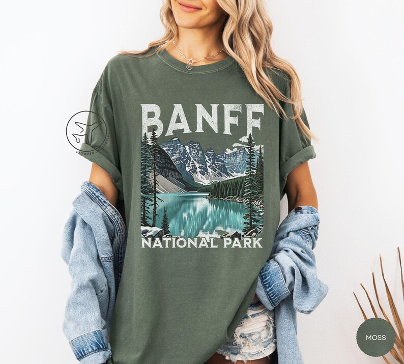 Może przedstawiać: Zielona koszulka w kolorze mchu z grafiką Parku Narodowego Banff. Projekt zawiera napis "BANFF" nad malowniczą ilustracją jeziora, g&oacute;r i drzew. Poniżej ilustracji znajdują się słowa "NATIONAL PARK".