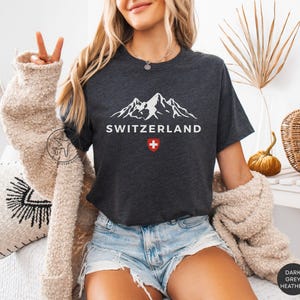 K&ouml;nnte beinhalten: Dunkelgraues meliertes T-Shirt mit wei&szlig;er Berg-Grafik, dem Wort "SWITZERLAND" und einem rot-wei&szlig;en Schweizer Kreuz. Das Shirt ist kurz&auml;rmlig und hat einen Rundhalsausschnitt. Es wird mit Jeansshorts und einer beigen Strickjacke getragen.