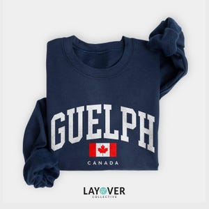 May include: A navy blue sweatshirt featuring the word "GUELPH" in white, bold lettering. Beneath this is a small Canadian flag and the word "CANADA". The garment is folded, with the Layover Collective logo visible at the bottom.