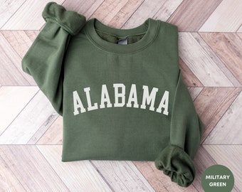 May include: A green sweatshirt with the word "ALABAMA" printed in white on the front. The sweatshirt is folded and laid on a wooden surface.