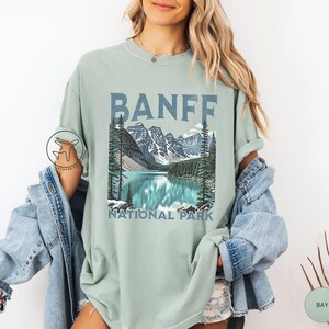 Może przedstawiać: Jasnozielona koszulka z napisem "BANFF" w niebieskich literach blokowych nad ilustracją krajobrazu jeziora i g&oacute;r. Pod ilustracją znajdują się słowa "NATIONAL PARK".