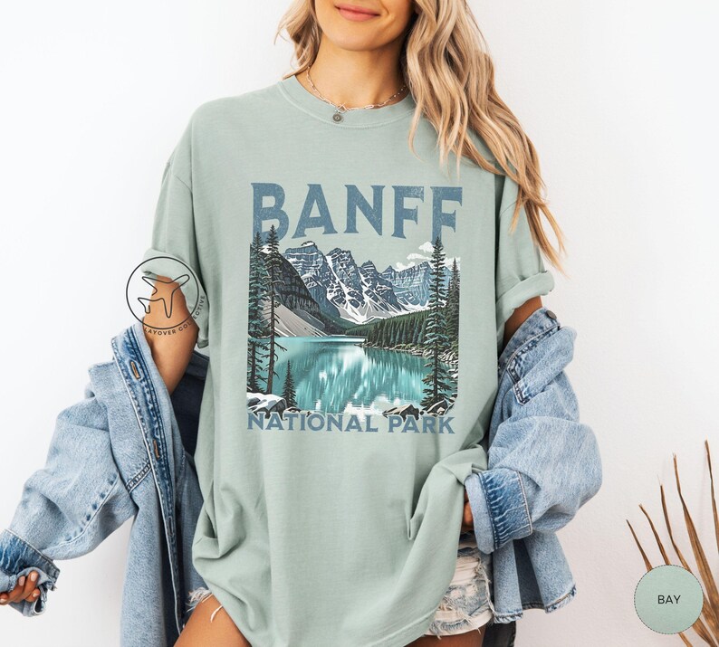 Może przedstawiać: Jasnozielona koszulka z napisem "BANFF" w niebieskich literach blokowych nad ilustracją krajobrazu jeziora i g&oacute;r. Pod ilustracją znajdują się słowa "NATIONAL PARK".