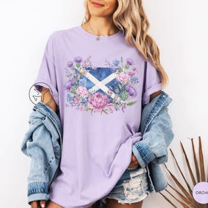 May include: A light purple t-shirt featuring a floral design around the Scottish flag. The design includes pink, purple, and blue flowers. The shirt has short sleeves and a relaxed fit.