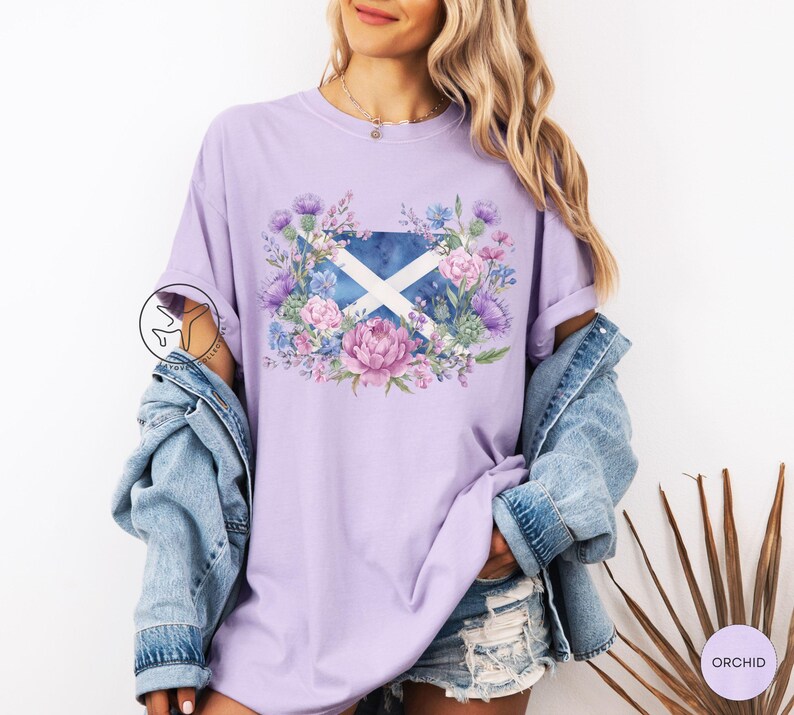 May include: A light purple t-shirt featuring a floral design around the Scottish flag. The design includes pink, purple, and blue flowers. The shirt has short sleeves and a relaxed fit.