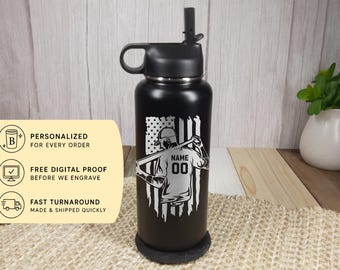 May include: A matte black insulated water bottle with a black lid and straw. The front features a white laser-engraved graphic of a baseball player holding a bat, positioned in front of a distressed American flag design with the text NAME and 00.