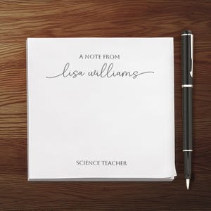 以下が含まれることがあります： 白いノートに「A note from lisa williams Science Teacher」という文字が印刷されています。黒いペンがノートの横に木製の表面に置かれています。