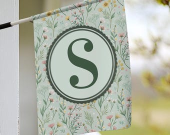 Könnte beinhalten: Ein grüner Buchstabe "S" in einem weißen Kreis mit einem gewellten Rand, auf grünem Hintergrund mit einem Blumenmuster aus weißen, rosa und gelben Blumen.