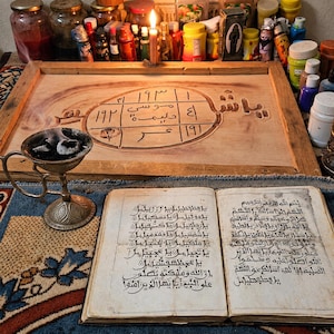 Könnte beinhalten: Ein aufgeschlagenes antikes Buch mit arabischer Schrift liegt vor einem Holzbrett, das ein zentrales ovales Design mit arabischen Ziffern und Text zeigt. Ein silberfarbenes Metall-Räuchergefäß mit glühender Kohle steht links daneben.
