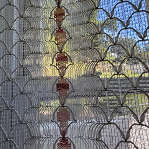 May include: A white sheer curtain featuring a repeating lattice pattern of interconnected loops. A vertical column of embroidered diamond-shaped motifs in gradient shades of orange and red runs down the center of the fabric, creating a textured design.