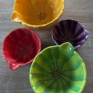 May include: Set of four colorful ceramic measuring bowls. The bowls are shaped like flowers in shades of yellow, red, purple, and green. Each bowl has a spout and a measurement label. The yellow bowl is labeled "1 cup".