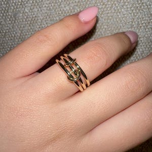 Könnte beinhalten: Ein goldener Ring mit einem Knotendesign. Der Ring besteht aus drei miteinander verbundenen Bändern.