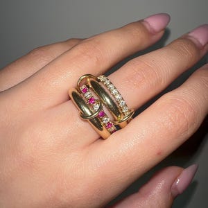 May include: A set of three gold rings with a delicate design. The top ring features a row of small pink gemstones, the middle ring has a row of small white gemstones, and the bottom ring is a plain gold band.