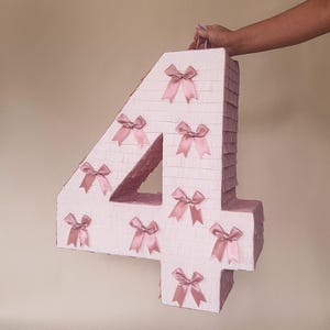 Puede incluir: Una piñata con forma de número cuatro, blanca y decorada con lazos rosas.