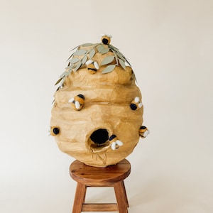 May include: A brown paper mache beehive with a small opening, decorated with green paper leaves and yellow and black felt bees. The beehive is sitting on a wooden stool.