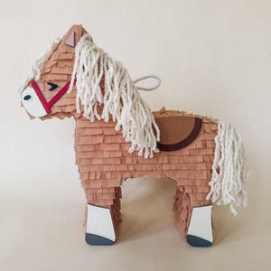 May include: A brown horse piñata with a white mane and tail. The piñata has a red bridle and a brown saddle. The horse's legs are white with blue hooves. A loop is attached to the top for hanging.