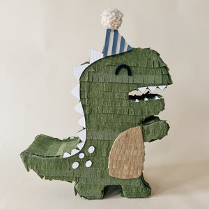 May include: Green dinosaur shaped pinata with a white pom-pom and blue and white striped party hat. The pinata has white polka dots and a brown tail.
