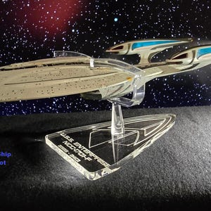 May include: A clear acrylic display stand for a model starship, featuring a triangular base engraved with the text U.S.S. ENTERPRISE NCC-1701-F and a curved support arm. The text Base Only - Ship and Stand Not Included is visible.
