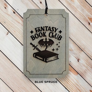 May include: A wooden sign with a light gray stain and the text "Fantasy Book Club" in black. A black silhouette of a dragon with wings spread and a sword is above a closed book. The text "Blue Spruce" is at the bottom of the sign.