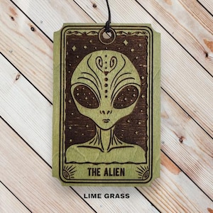 May include: A lime green and brown wooden plaque featuring an alien design. The alien has large oval eyes and a detailed face. The text "THE ALIEN" is at the bottom, with "LIME GRASS" below it. A black string is attached for hanging.