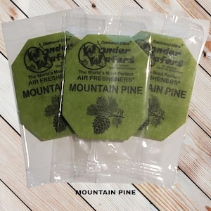 May include: Three green air fresheners in clear packaging. Each air freshener has the text "MOUNTAIN PINE" and a pine cone illustration. The packaging also includes the text "Wonder Wafers" and "The World's Most Perfect Air Fresheners."