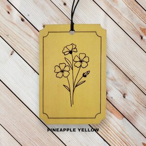 May include: A rectangular, pineapple yellow wooden tag with a black floral illustration. The tag has a black border and a hole at the top for hanging. The text "PINEAPPLE YELLOW" is at the bottom.