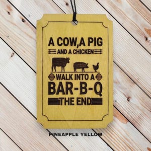 May include: A yellow wooden sign with the text "A cow, a pig and a chicken walk into a bar-b-q the end". The sign has a black string attached to the top for hanging.