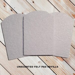 May include: Three light gray, unscented felt pad refills with a glittery texture. The pads have a rounded top and a rectangular base. The text "UNSCENTED FELT PAD REFILLS" is printed in black at the bottom of the image.