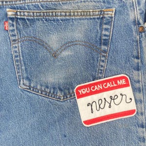 May include: Close-up of a light blue denim jeans pocket with a red and white patch that reads "YOU CAN CALL ME" above the word "never" in black script. The jeans have a classic five-pocket design.