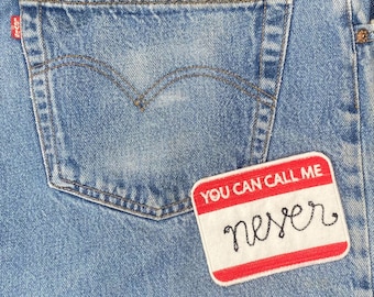 May include: Close-up of a light blue denim jeans pocket with a red and white patch that reads "YOU CAN CALL ME" above the word "never" in black script. The jeans have a classic five-pocket design.