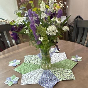 May include: A green and purple quilted table runner with a starburst design. The runner is on a wooden table with a vase of purple and white flowers in the center.