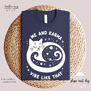 K&ouml;nnte beinhalten: Ein Unisex-T-Shirt in der Farbe Blau-Melange mit einem wei&szlig;en Motiv eines Katzen, das sich in einem Kreis mit Sternen und Planeten im Inneren zusammenrollt. Der Text "Me and Karma Vibe Like That" ist unter der Katze aufgedruckt.