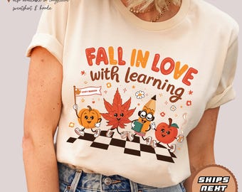 May include: Cream-colored unisex t-shirt with the text "FALL IN LOVE with learning" in orange and red. The graphic features cartoon characters of a pumpkin, leaf, pencil, and apple. The shirt also has the text "COZY SEASON".
