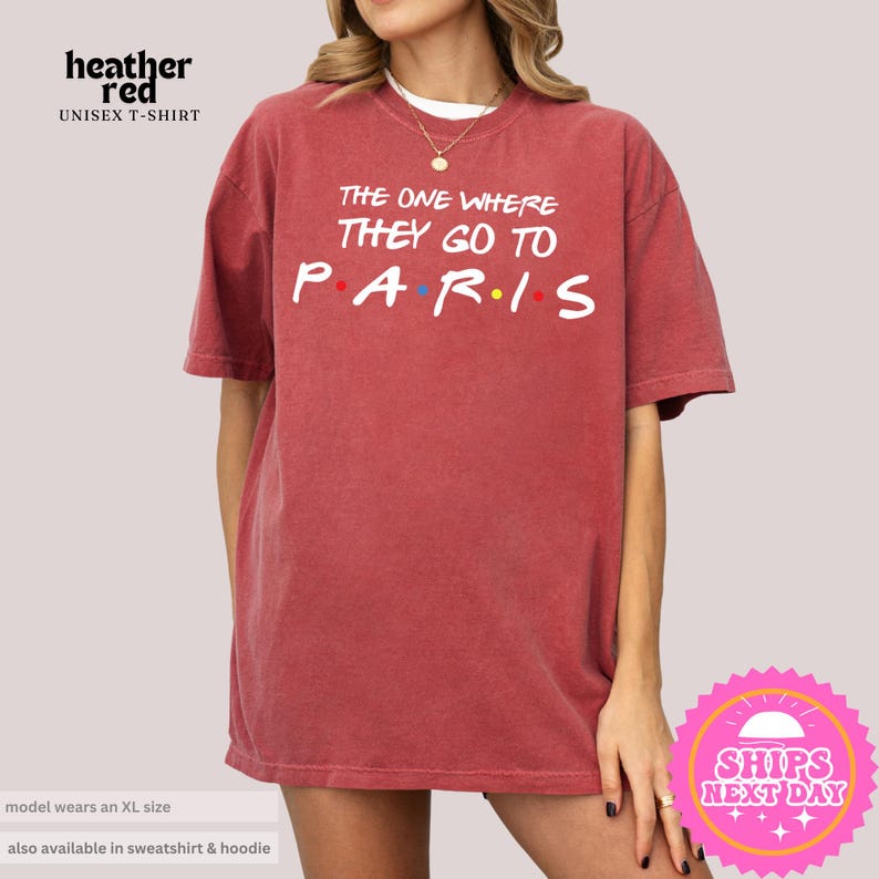 May include: Red unisex t-shirt with the text "The One Where They Go To Paris" in white with colorful dots. The shirt is a relaxed fit and has a distressed look.