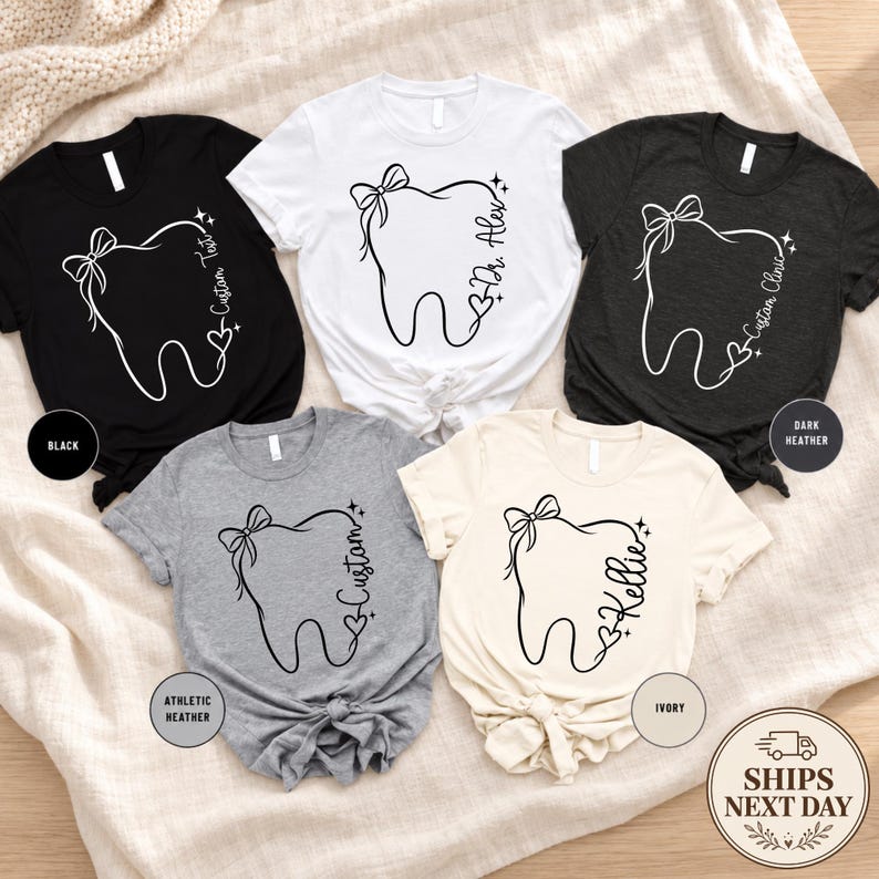 May include: Five short-sleeved t-shirts in black, white, dark heather, athletic heather, and ivory. Each shirt features a line art graphic of a tooth with a bow, a heart, and custom text. The shirts are arranged on a textured fabric surface.
