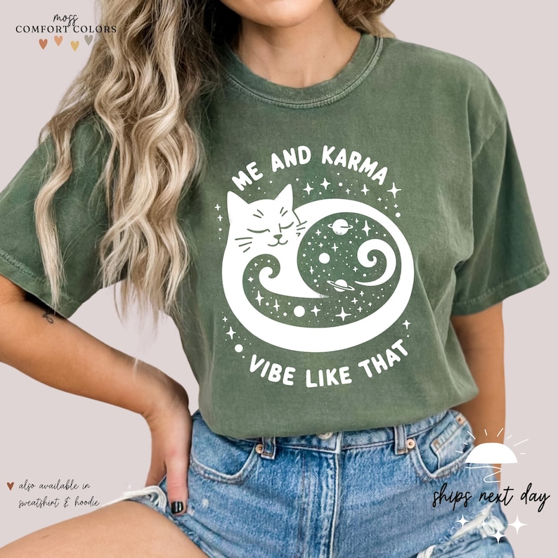 K&ouml;nnte beinhalten: Ein gr&uuml;nes T-Shirt mit einer wei&szlig;en Grafik eines Katers, der sich in einem Kreis mit Sternen und Planeten darin zusammenrollt. Der Text "Me and Karma Vibe Like That" ist unter dem Kater aufgedruckt.