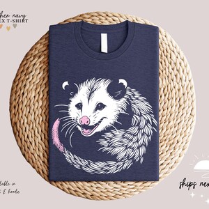 Puede incluir: Camiseta unisex azul marino jaspeado con un dise&ntilde;o de zarig&uuml;eya en blanco y negro, con nariz y cola rosas. El texto "heather navy" y "UNISEX T-SHIRT" est&aacute;n en la parte superior. Tambi&eacute;n disponible en sudadera y sudadera con capucha.