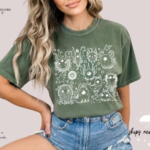 May include: A green t-shirt with a white graphic print of a group of cartoon monsters with large eyes. The graphic is surrounded by stars and other celestial elements. The text "moss & comfort colors" is in the top left corner of the image.