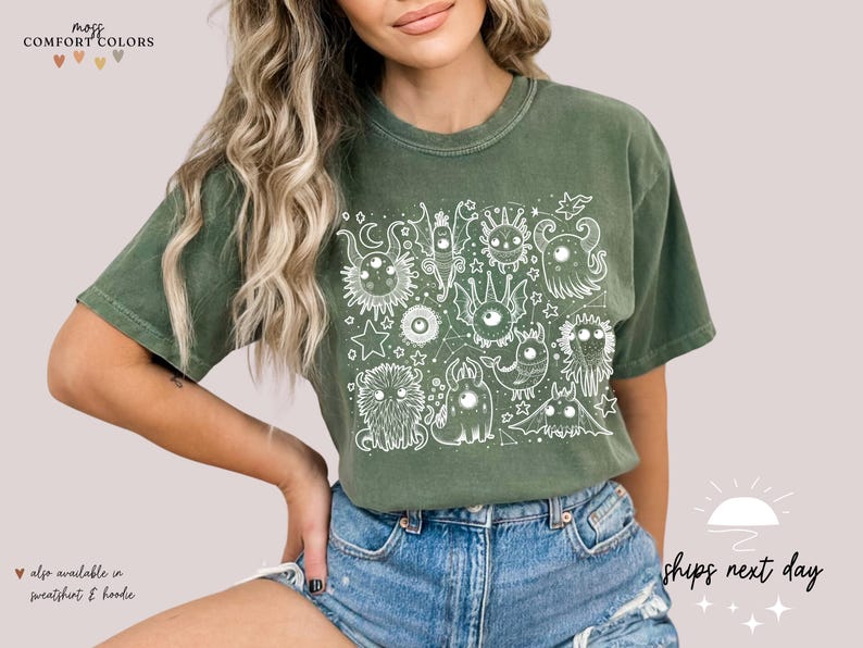 May include: A green t-shirt with a white graphic print of a group of cartoon monsters with large eyes. The graphic is surrounded by stars and other celestial elements. The text "moss & comfort colors" is in the top left corner of the image.