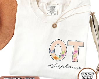 May include: Cream-colored t-shirt with the letters "OT" in a floral design, with the name "Stephanie" below. A gold necklace with a pendant rests on the shirt. The shirt also has the text "MORE STYLES + COLORS" and "SHIPS NEXT DAY".