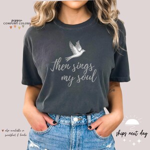 Puede incluir: Una camiseta gris oscuro con una paloma blanca y el texto "Then sings my soul" impreso en la parte delantera.