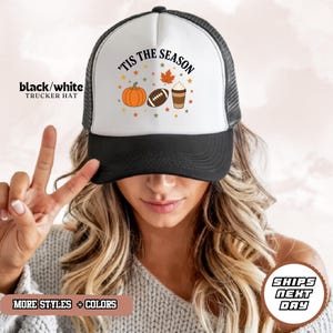 May include: A black and white trucker hat featuring a white front panel with the text 'TIS THE SEASON in black lettering, accompanied by a graphic print of an orange pumpkin, a brown football, and a coffee cup surrounded by small autumn leaves.