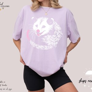 Puede incluir: Camiseta lila claro con un dise&ntilde;o de zarig&uuml;eya blanca y lengua rosa. La zarig&uuml;eya est&aacute; rodeada por un c&iacute;rculo rosa. El texto "orchid COMFORT COLORS" est&aacute; en la parte superior izquierda. El texto "ships next day" est&aacute; en la parte inferior derecha.