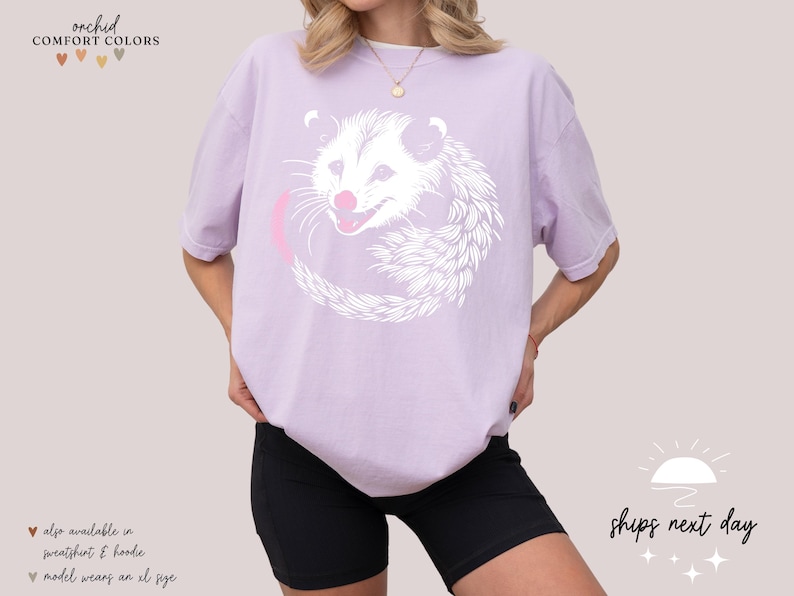 Puede incluir: Camiseta lila claro con un dise&ntilde;o de zarig&uuml;eya blanca y lengua rosa. La zarig&uuml;eya est&aacute; rodeada por un c&iacute;rculo rosa. El texto "orchid COMFORT COLORS" est&aacute; en la parte superior izquierda. El texto "ships next day" est&aacute; en la parte inferior derecha.