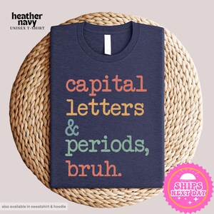 K&ouml;nnte beinhalten: Ein T-Shirt in Marineblau mit dem Text "capital letters & periods, bruh." in Regenbogenfarben.