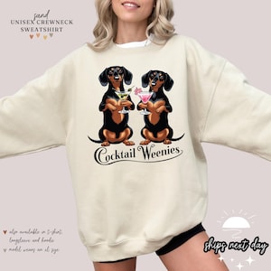 May include: Sand-colored unisex crewneck sweatshirt featuring a graphic of two dachshunds holding cocktails. The phrase "Cocktail Weenies" is written below the dogs. The sweatshirt also says "ships next day".