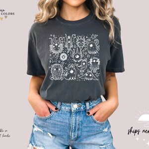 May include: A dark gray t-shirt with a white graphic print of a group of cartoon monsters. The shirt has a crew neck and short sleeves. The text "pepper COMFORT COLORS" is visible at the top of the image.