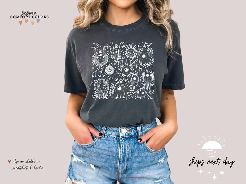May include: A dark gray t-shirt with a white graphic print of a group of cartoon monsters. The shirt has a crew neck and short sleeves. The text "pepper COMFORT COLORS" is visible at the top of the image.