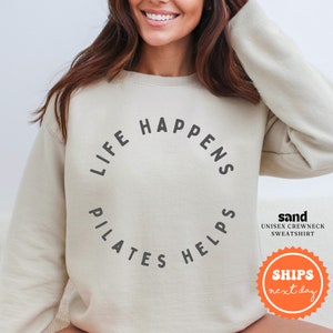 May include: A light beige sweatshirt with the text "Life Happens Pilates Helps" printed in a circle on the front. The sweatshirt is a unisex crewneck style.