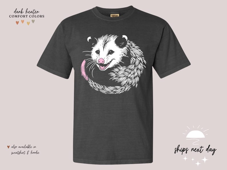 Puede incluir: Camiseta gris oscuro con una ilustraci&oacute;n de zarig&uuml;eya blanca, nariz y cola rosas. El texto "COMFORT COLORS" est&aacute; encima de la camiseta. Tambi&eacute;n disponible en sudadera y sudadera con capucha. El texto "ships next day" tambi&eacute;n es visible.